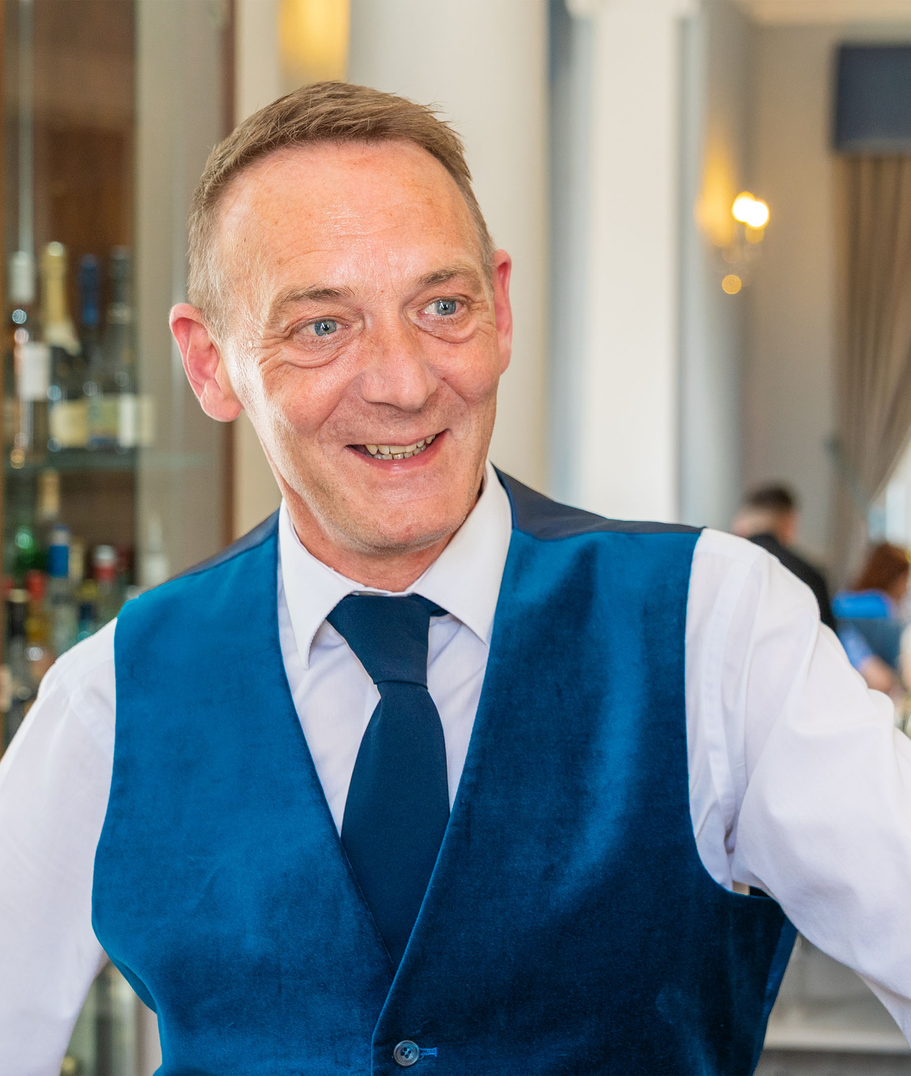 A man wearing a white shirt, blue waistcoat and blue tie standing over a bar serving residents