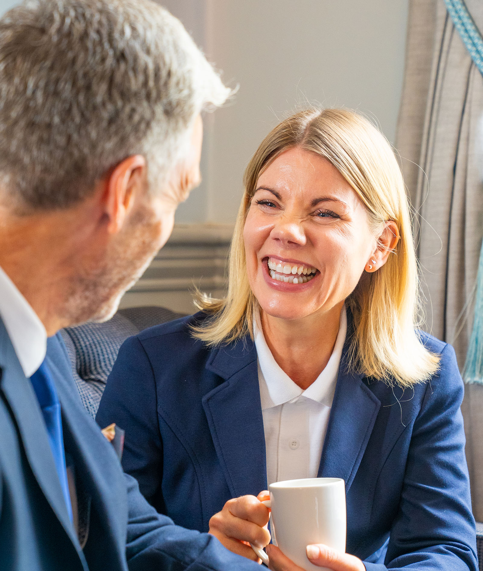 A woman with short blonde hair holding a mug of tea or coffee having a conversation with a male colleague and laughing