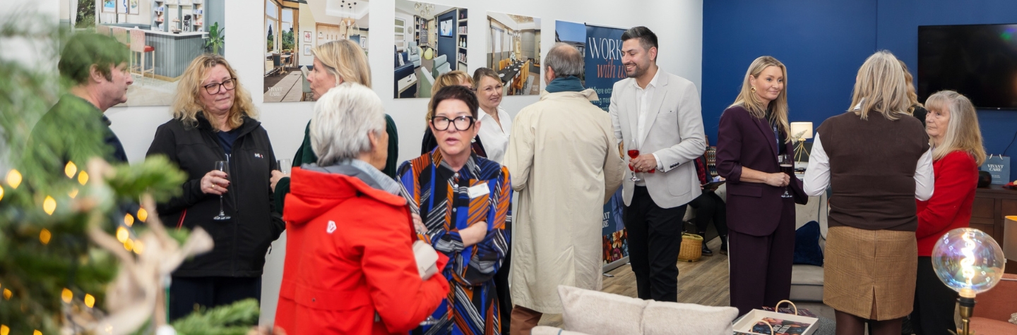  group of people gathered in a modern indoor setting, engaging in conversation during a social or networking event. The room features a blue accent wall, framed interior design photographs on the white wall, and a patterned armchair in the foreground. A decorated Christmas tree is partially visible on the left, and some attendees are holding drinks.