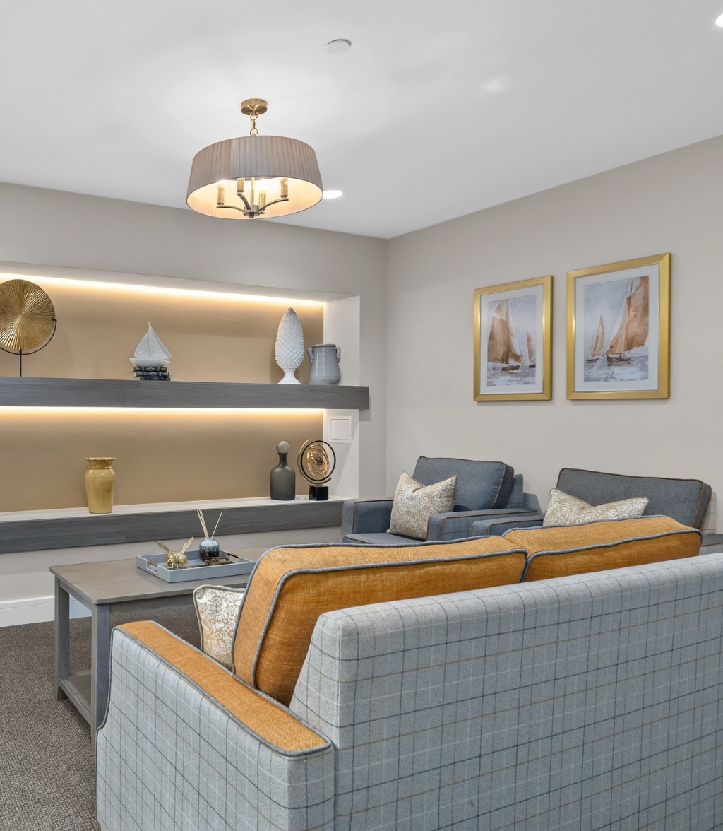 Stylish care home lounge with plaid sofas, blue armchairs, a coffee table, warm backlit shelving displaying decorative ornaments, and framed sailboat artwork on the wall